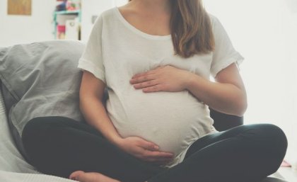 A heavily pregnant woman sits cross-legged with her arms protectively around the top and bottom of her belly.